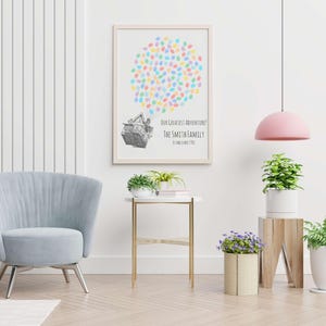 Puede incluir: Una impresi&oacute;n enmarcada con un marco de madera clara. La impresi&oacute;n presenta un grupo de huellas dactilares coloridas sobre un dibujo detallado de una casa. El texto debajo dice "OUR GREATEST ADVENTURE! THE SMITH FAMILY". Tambi&eacute;n se ven una silla azul claro y una mesa auxiliar.