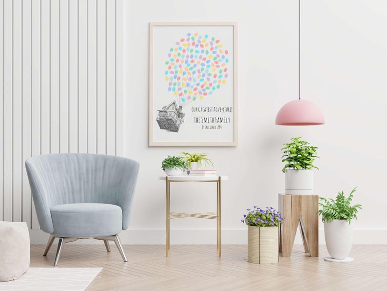 Puede incluir: Una impresi&oacute;n enmarcada con un marco de madera clara. La impresi&oacute;n presenta un grupo de huellas dactilares coloridas sobre un dibujo detallado de una casa. El texto debajo dice "OUR GREATEST ADVENTURE! THE SMITH FAMILY". Tambi&eacute;n se ven una silla azul claro y una mesa auxiliar.