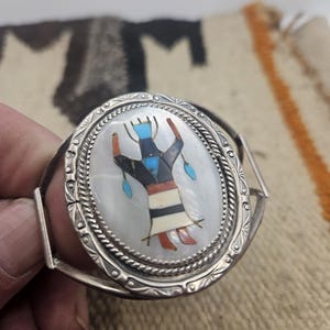 May include: Oval silver bracelet with a detailed inlaid design. The centerpiece features a figure with a black, red, and white pattern, set against a mother-of-pearl background. The bracelet has a decorative border.