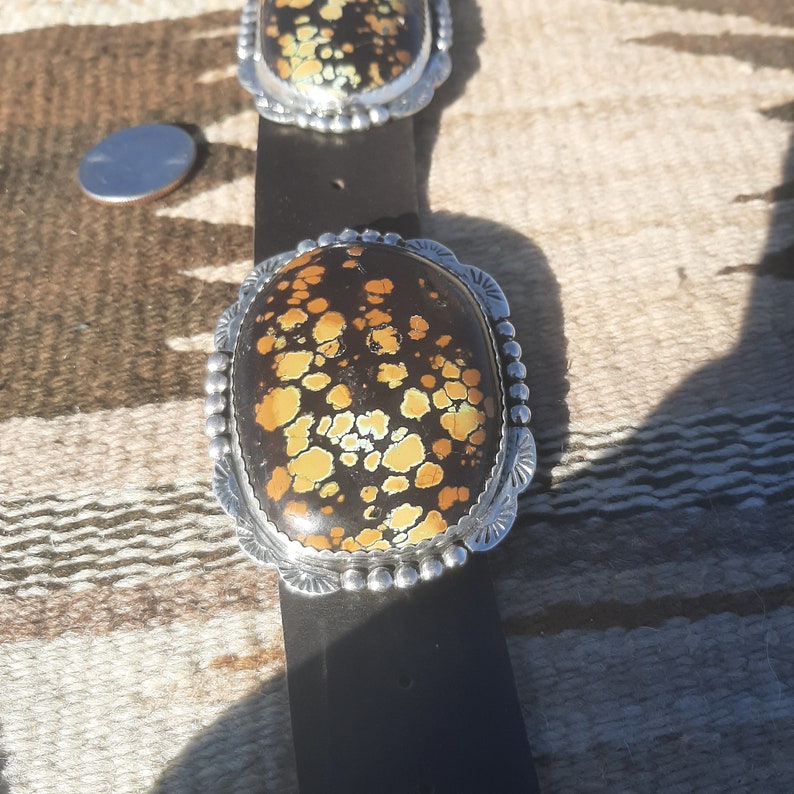 May include: A brown leather belt with a silver buckle featuring a large oval-shaped stone with a brown and yellow mottled pattern.