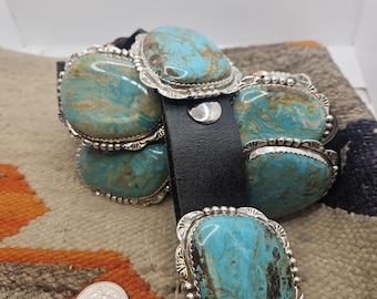 May include: A black leather cuff bracelet featuring five large, oval-shaped turquoise stones with brown matrix patterns. Each stone is set in a scalloped silver bezel with beaded detailing. A silver snap closure secures the band, shown with a US coin.