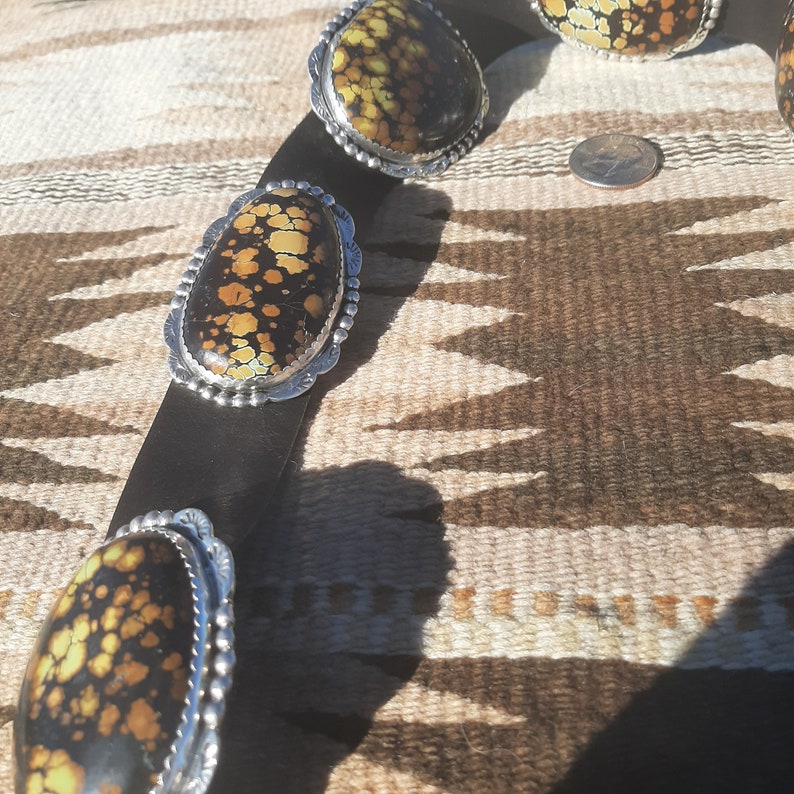 May include: A black leather belt with silver oval-shaped buckles set with yellow and black patterned stones.