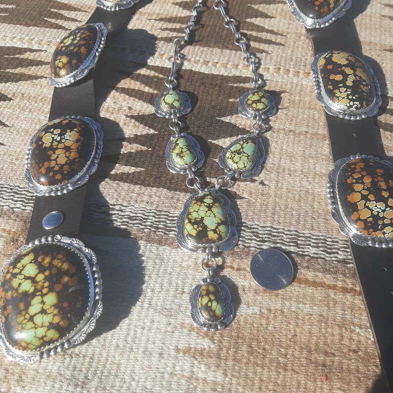 May include: A turquoise and silver necklace and belt set with a southwestern design. The necklace has five turquoise stones and the belt has four turquoise stones.