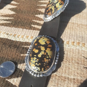 May include: A brown leather belt with two oval-shaped silver belt buckles. Each buckle features a dark brown stone with yellow and green splotches.