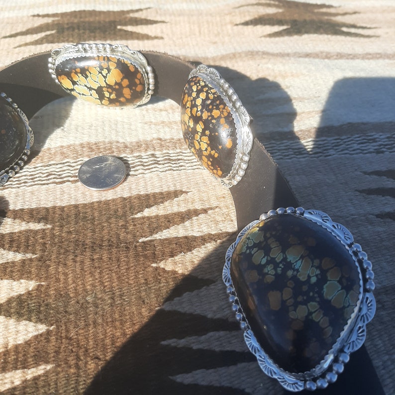 May include: A leather belt with four silver and turquoise concho buckles. The belt is on a brown and white patterned surface.