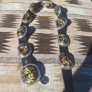 May include: A brown leather belt with silver oval-shaped buckles set with green, brown, and yellow turquoise stones.