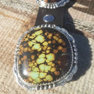 May include: A silver belt buckle with a large, oval-shaped turquoise stone. The stone has a black and brown matrix with green and yellow veins.