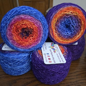 May include: A collection of colorful yarn skeins in shades of blue, purple, orange, and pink. The yarn is arranged in a gradient pattern, with each skein displaying a unique color transition. Each skein has a tag with the text "UPNORTH YARNS - one skein at a time".