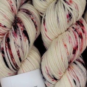 May include: Close-up of two skeins of white yarn with red, pink, and black speckles. The yarn is labeled "Cardinals in Winter" and is made of Superwash 100% Merino wool. The yarn is Worsted Weight.