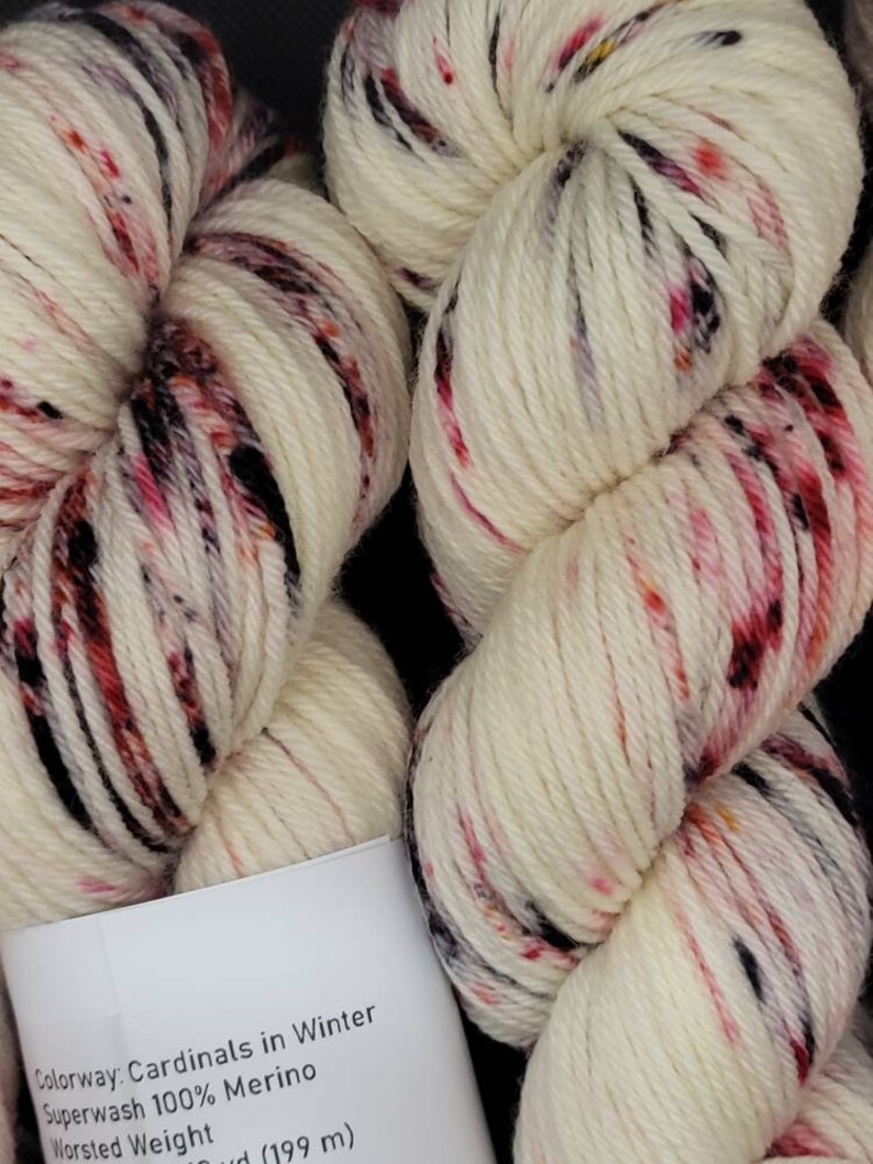 May include: Close-up of two skeins of white yarn with red, pink, and black speckles. The yarn is labeled "Cardinals in Winter" and is made of Superwash 100% Merino wool. The yarn is Worsted Weight.