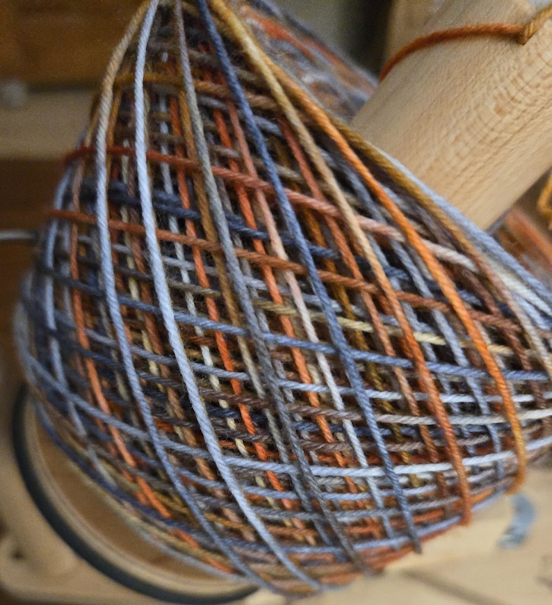 Puede incluir: Un ovillo de hilo multicolor enrollado en un huso de madera, con una mezcla de colores que incluye tonos naranja, &oacute;xido, marr&oacute;n, azul pizarra y gris claro. El hilo est&aacute; enrollado en un patr&oacute;n entrecruzado alrededor del n&uacute;cleo de madera.