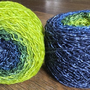 May include: Two skeins of yarn sit side-by-side. One is a ball of yarn with a gradient of green and blue hues. The other is a cylindrical skein of dark blue yarn with white flecks, revealing green yarn at the center. The yarn is on a wooden surface.