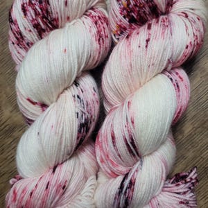 May include: Two skeins of white yarn with vibrant red and black speckles. The yarn is tightly wound, showcasing the texture and color variations. Ideal for knitting or crochet projects.