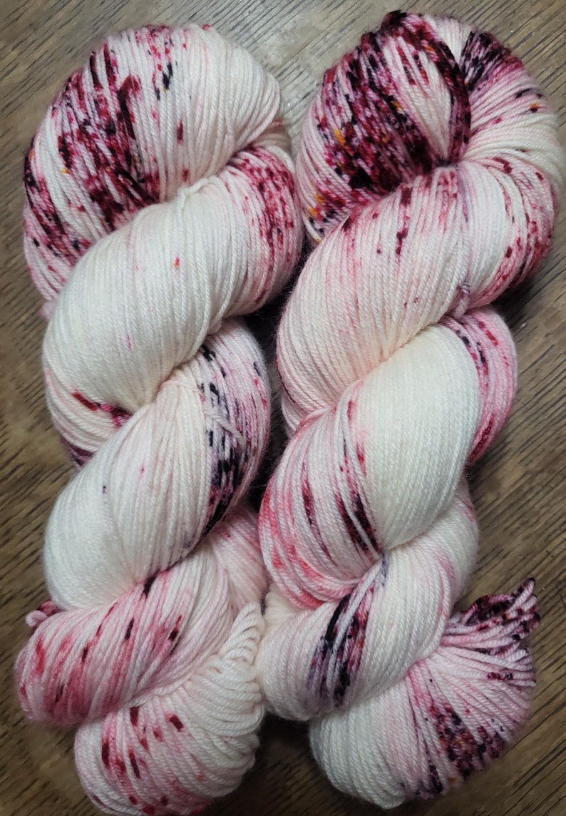 May include: Two skeins of white yarn with vibrant red and black speckles. The yarn is tightly wound, showcasing the texture and color variations. Ideal for knitting or crochet projects.