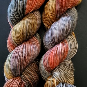 Puede incluir: Dos madejas de hilo te&ntilde;ido a mano con una paleta de colores variados que incluye naranja quemado, amarillo mostaza, verde salvia y gris pizarra. Las hebras de fibra trenzadas muestran una compleja mezcla de tonos tierra en toda su longitud.