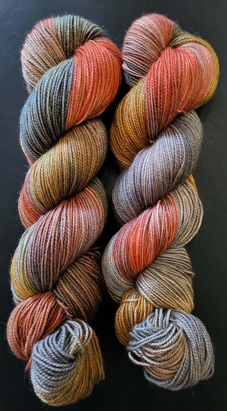 Puede incluir: Dos madejas de hilo te&ntilde;ido a mano con una paleta de colores variados que incluye naranja quemado, amarillo mostaza, verde salvia y gris pizarra. Las hebras de fibra trenzadas muestran una compleja mezcla de tonos tierra en toda su longitud.