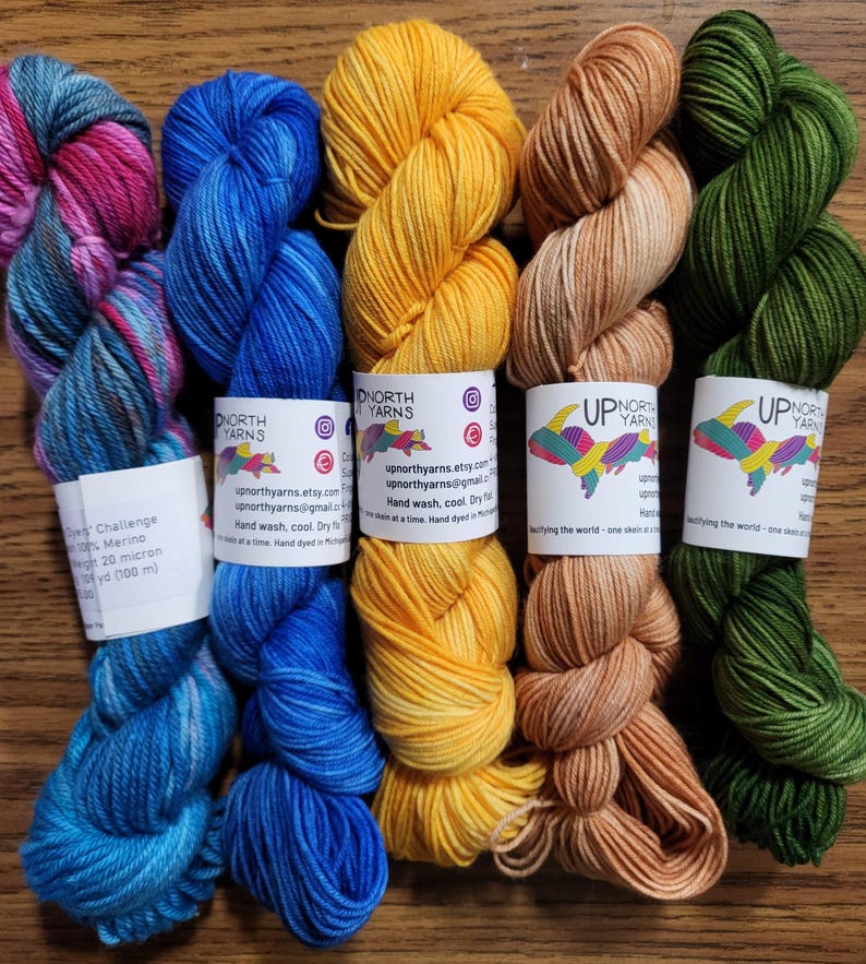 May include: Five skeins of yarn arranged side-by-side, featuring colors of variegated pink and blue, solid bright blue, solid yellow, solid light tan, and solid dark green. Each skein has a white paper label with the text Up North Yarns.