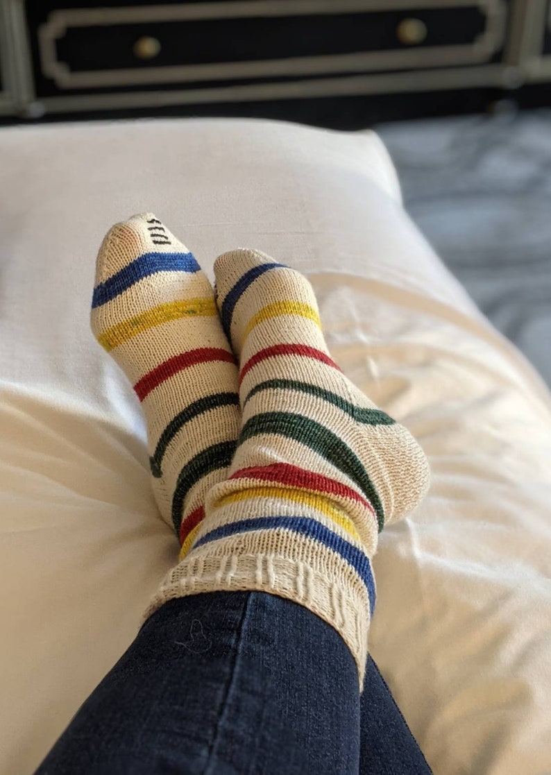 Puede incluir: Un par de calcetines a rayas con rayas rojas, amarillas, azules y verdes sobre un fondo blanco. Los calcetines se llevan puestos en los pies de una persona y la persona lleva pantalones vaqueros azules.