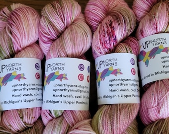 Puede incluir: Varios ovillos de hilo teñido a mano en tonos rosa, verde y crema. Cada ovillo tiene una etiqueta con el texto "UP NORTH YARNS" e iconos de redes sociales. El hilo está dispuesto sobre una superficie de madera.