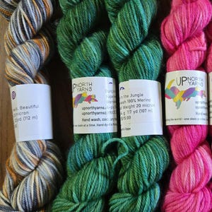 May include: Four skeins of Up North Yarns wool yarn. The first is grey, white, and orange variegated. The second and third are solid green. The fourth is bright pink. Each skein has a white paper label with text including 100% Merino wool.