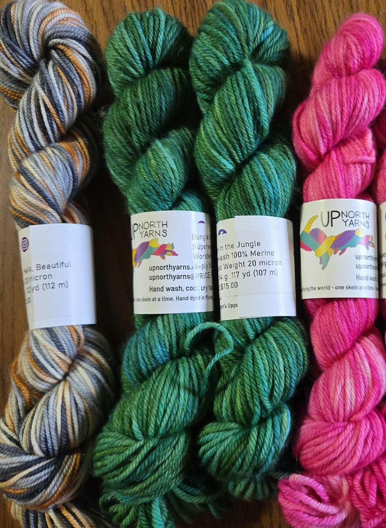 May include: Four skeins of Up North Yarns wool yarn. The first is grey, white, and orange variegated. The second and third are solid green. The fourth is bright pink. Each skein has a white paper label with text including 100% Merino wool.