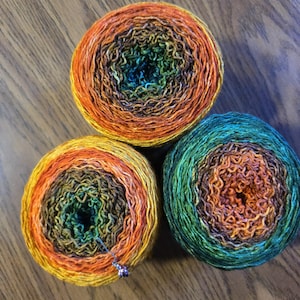 May include: Three colorful yarn cakes in shades of orange, yellow, green, and blue sit on a wooden surface. The yarn is wound in concentric circles, creating a gradient effect. A small beaded charm is attached to one of the yarn cakes.