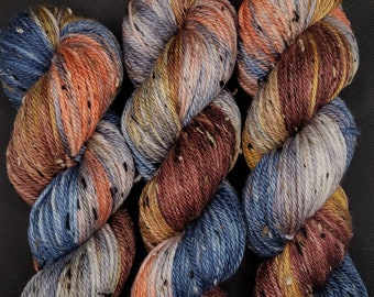 Puede incluir: Tres madejas de hilo teñido a mano con una paleta de colores variados en azul polvoriento, naranja quemado, burdeos intenso y amarillo dorado. El hilo tiene una construcción trenzada con pequeñas motas de color marrón oscuro y blanco.