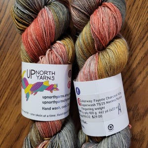 Puede incluir: Dos madejas de hilo te&ntilde;ido a mano de grosor fingering en un color variado de gris carb&oacute;n, naranja apagado y amarillo mostaza. Las etiquetas de papel blanco muestran el logotipo de Up North Yarns y detalles sobre la mezcla 75/25 merino/nylon.