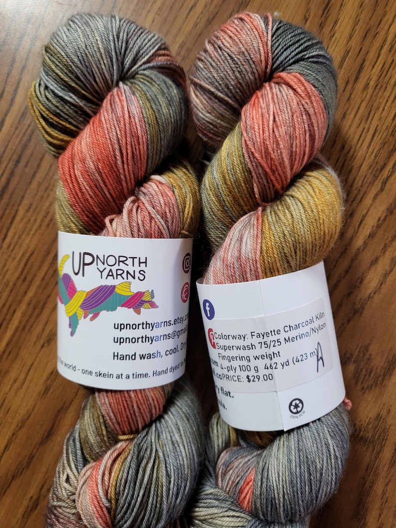 Puede incluir: Dos madejas de hilo te&ntilde;ido a mano de grosor fingering en un color variado de gris carb&oacute;n, naranja apagado y amarillo mostaza. Las etiquetas de papel blanco muestran el logotipo de Up North Yarns y detalles sobre la mezcla 75/25 merino/nylon.