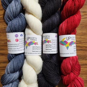 May include: Four skeins of fingering weight yarn arranged side-by-side, featuring colors of heathered blue, cream, charcoal gray, and vibrant red. Each skein has a white paper label with the text Up North Yarns and a colorful fish graphic.