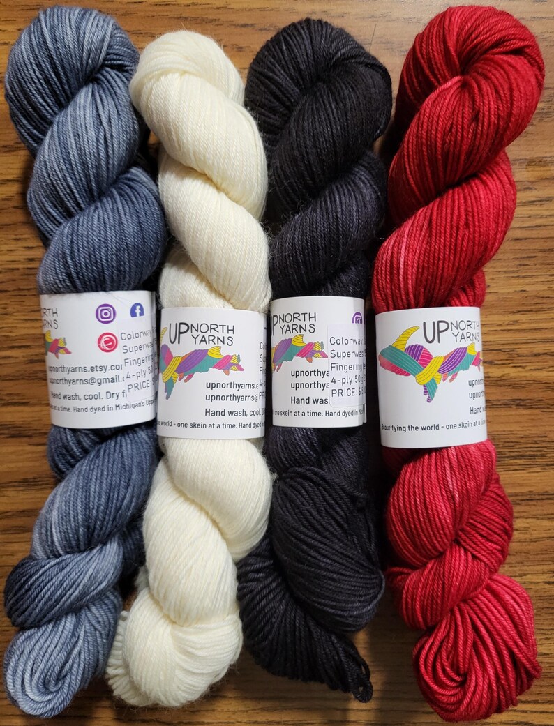 May include: Four skeins of fingering weight yarn arranged side-by-side, featuring colors of heathered blue, cream, charcoal gray, and vibrant red. Each skein has a white paper label with the text Up North Yarns and a colorful fish graphic.
