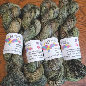 May include: Four skeins of hand-dyed yarn in variegated shades of olive green, brown, and charcoal gray. Each skein features a white paper label with the text Up North Yarns, social media icons, and contact information, noting it is hand-dyed in Michigan.