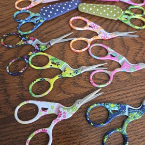 May include: A collection of small metal embroidery scissors featuring various colorful floral and polka dot patterns on the handles and blades. Some scissors are paired with matching fabric-covered protective sheaths in matching designs.