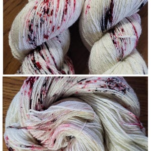 May include: Close-up of two skeins of white yarn with red and black speckles and silver metallic thread. The yarn is tightly wound, showing the texture and color variations. Ideal for knitting or crochet projects.