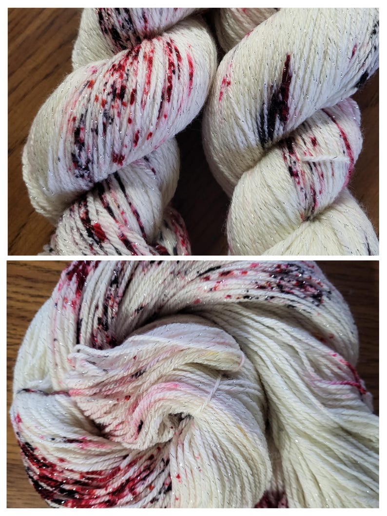 May include: Close-up of two skeins of white yarn with red and black speckles and silver metallic thread. The yarn is tightly wound, showing the texture and color variations. Ideal for knitting or crochet projects.