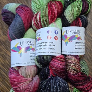 May include: Three skeins of hand-dyed yarn in shades of red, green, and black with silver glitter. The yarn is labeled "Up North Yarns" and includes contact information and the phrase "Hand wash, cool dry flat."