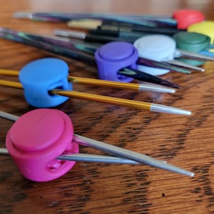 May include: A close-up of colorful knitting needle holders. The holders are made of plastic and have a circular shape. The needles are made of metal and are in various colors.