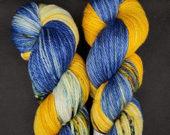 Puede incluir: Dos madejas de hilo teñido a mano con una paleta de colores variados en amarillo vibrante, azul intenso y blanco crema. Cada madeja tiene una etiqueta de papel blanco con el texto Up North Yarns impreso en letras negras.