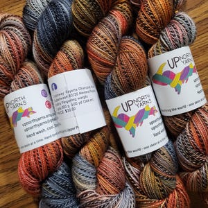 Puede incluir: Cuatro madejas de hilo te&ntilde;ido a mano de peso fingering con una paleta de colores variados en tonos carb&oacute;n, naranja quemado y azul apagado. Cada madeja est&aacute; envuelta en una etiqueta de papel blanco con el logotipo de Up North Yarns y detalles.
