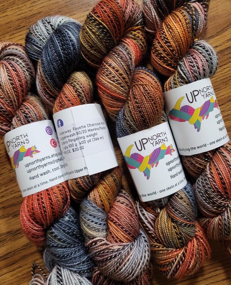 Puede incluir: Cuatro madejas de hilo te&ntilde;ido a mano de peso fingering con una paleta de colores variados en tonos carb&oacute;n, naranja quemado y azul apagado. Cada madeja est&aacute; envuelta en una etiqueta de papel blanco con el logotipo de Up North Yarns y detalles.