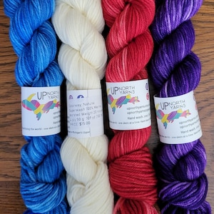 May include: Four skeins of Up North Yarns wool yarn in blue, natural white, red, and purple. Each skein features a white paper label with the brand logo, text, and care instructions. The yarn is twisted into a long, soft, textured bundle.