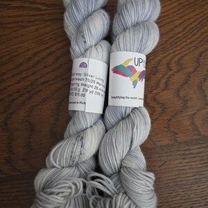 May include: Two skeins of light gray and white variegated yarn with a label reading Up North Yarns, Silver Lining, Superwash 75/25 Merino, Fingering Weight, 50 g, 218 yd, and Beautifying the world - one skein at a time.