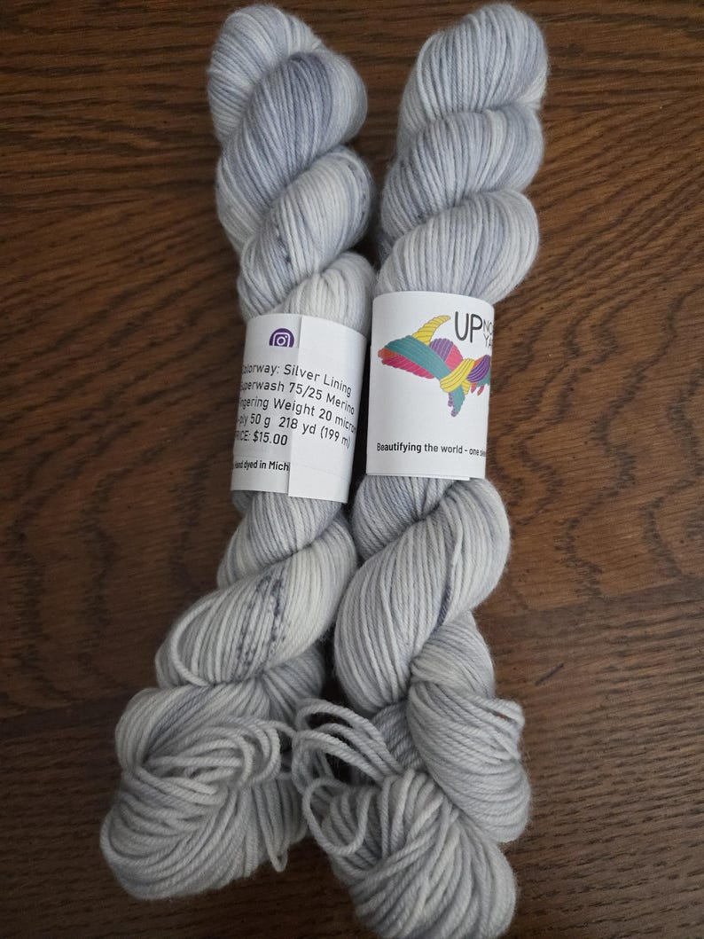 May include: Two skeins of light gray and white variegated yarn with a label reading Up North Yarns, Silver Lining, Superwash 75/25 Merino, Fingering Weight, 50 g, 218 yd, and Beautifying the world - one skein at a time.