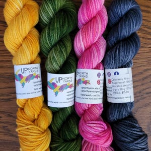May include: Four skeins of hand-dyed yarn in yellow, forest green, bright pink, and charcoal gray. Each skein features a white paper label with the Up North Yarns logo, contact information, and care instructions for 50g of 4-ply wool.