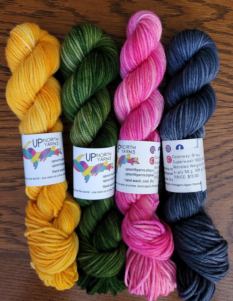 May include: Four skeins of hand-dyed yarn in yellow, forest green, bright pink, and charcoal gray. Each skein features a white paper label with the Up North Yarns logo, contact information, and care instructions for 50g of 4-ply wool.