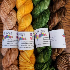 May include: Four skeins of hand-dyed yarn arranged side-by-side in shades of light tan, bright yellow, forest green, and dark brown. Each skein features a white paper label with the Up North Yarns logo, contact information, and care instructions.