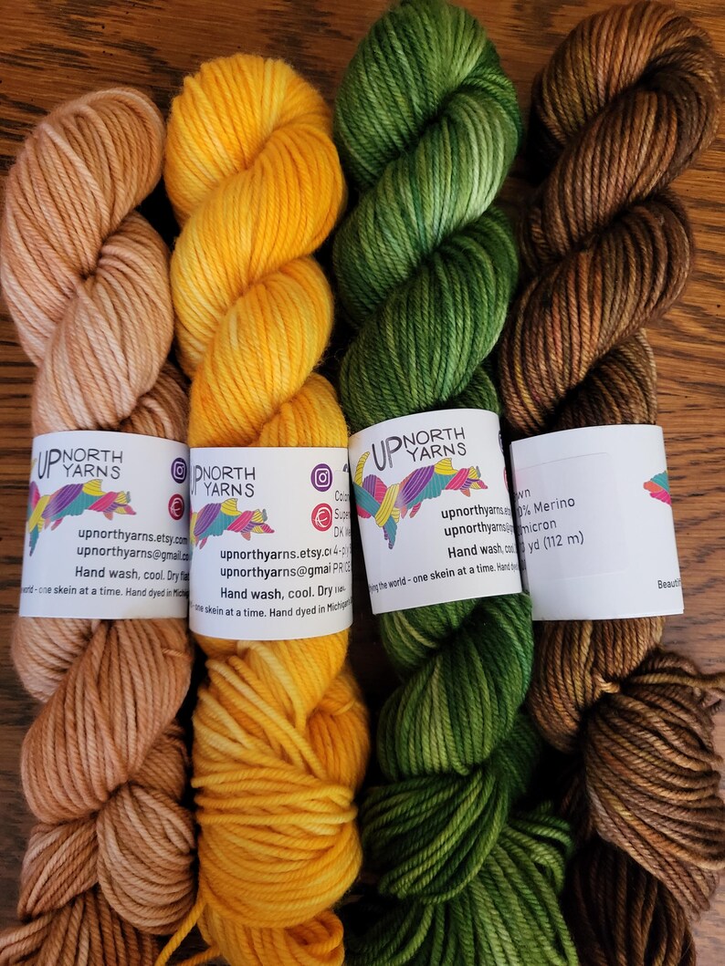 May include: Four skeins of hand-dyed yarn arranged side-by-side in shades of light tan, bright yellow, forest green, and dark brown. Each skein features a white paper label with the Up North Yarns logo, contact information, and care instructions.