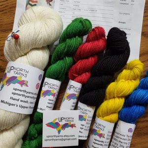Puede incluir: Surtido de ovillos de lana de colores en verde, rojo, negro, amarillo, azul y blanco. La lana est&aacute; etiquetada con "Up North Yarns" e incluye informaci&oacute;n sobre la composici&oacute;n de la lana, su peso y las instrucciones de cuidado. La lana es adecuada para proyectos de tejido o ganchillo.