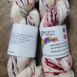 May include: Two skeins of white yarn with red and black speckles. The yarn is labeled "Cardinals in Winter" and is 100% Merino wool. Each skein weighs 100 grams and has 246 yards (225 m) of yarn.