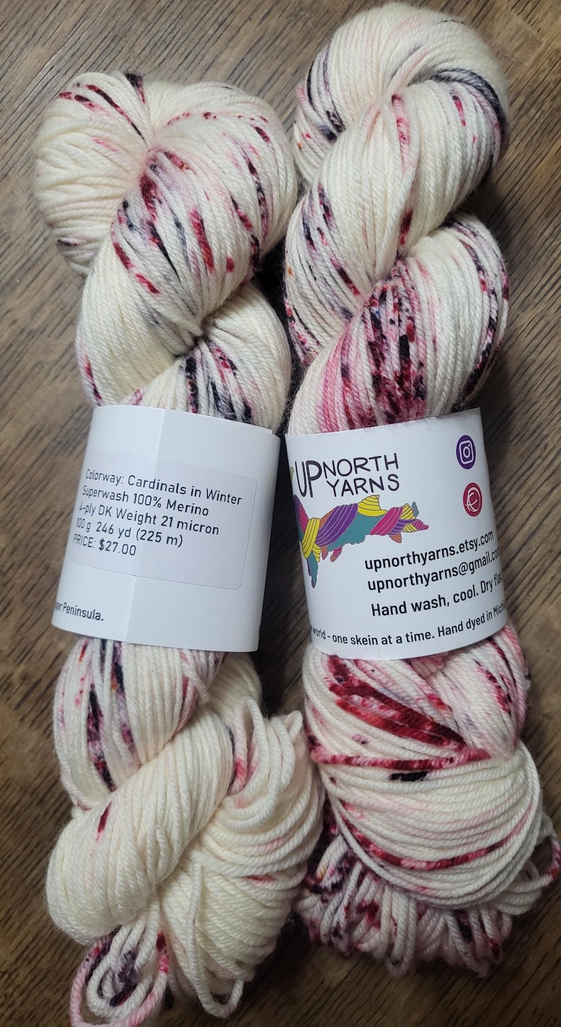 May include: Two skeins of white yarn with red and black speckles. The yarn is labeled "Cardinals in Winter" and is 100% Merino wool. Each skein weighs 100 grams and has 246 yards (225 m) of yarn.